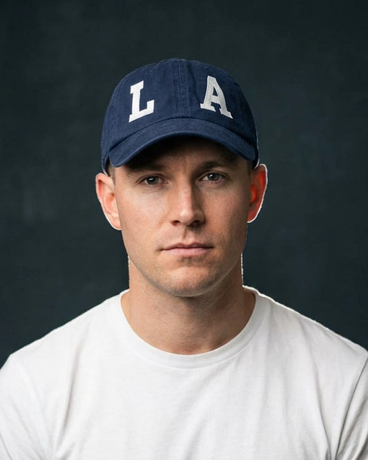 Male model wearing a navy blue dad cap with “LA” embroidery, photographed in a clean studio setup against a dark background.