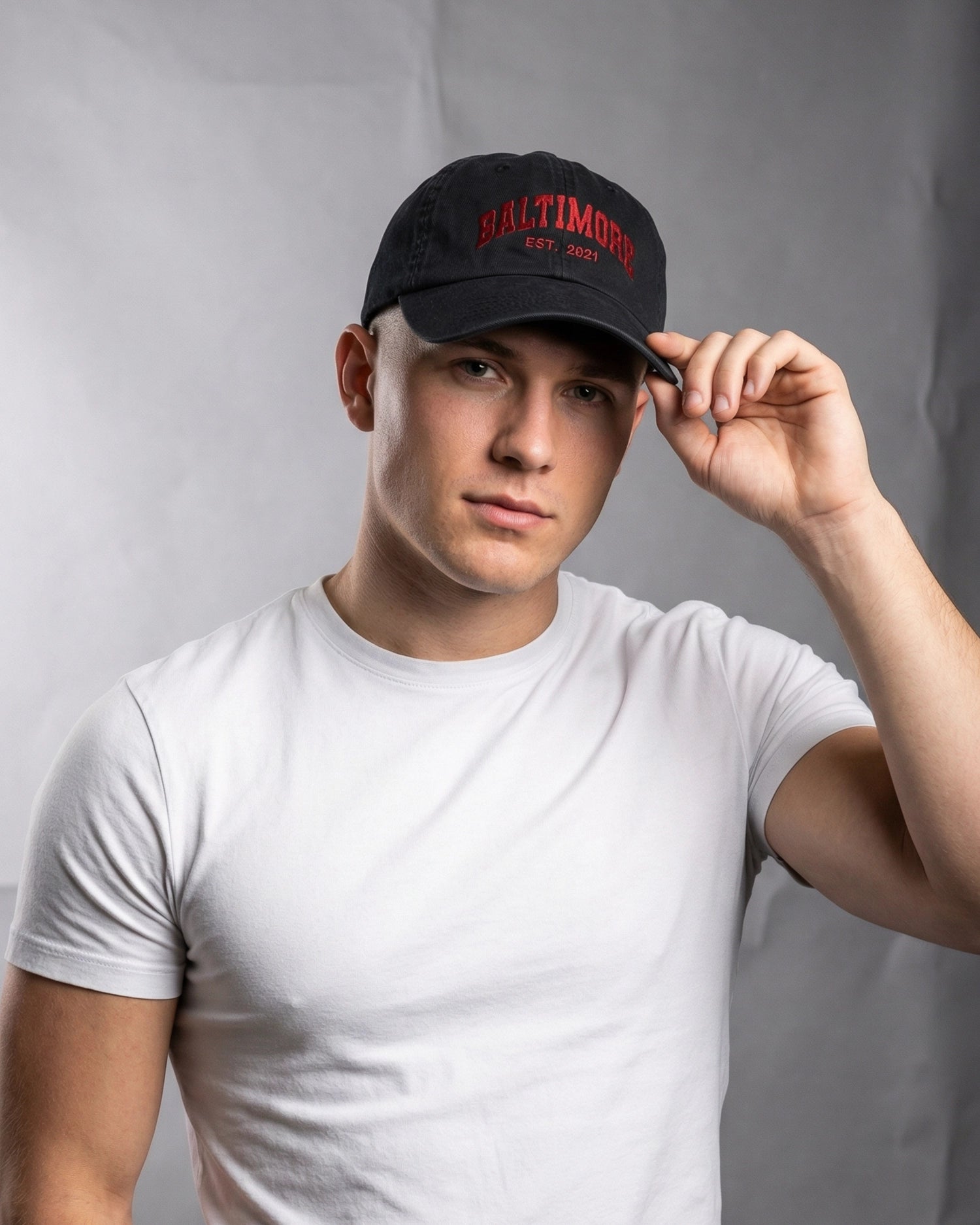 Man wearing the 999TEE Baltimore black dad cap with embroidered detailing, paired with a white t-shirt, against a neutral grey background.