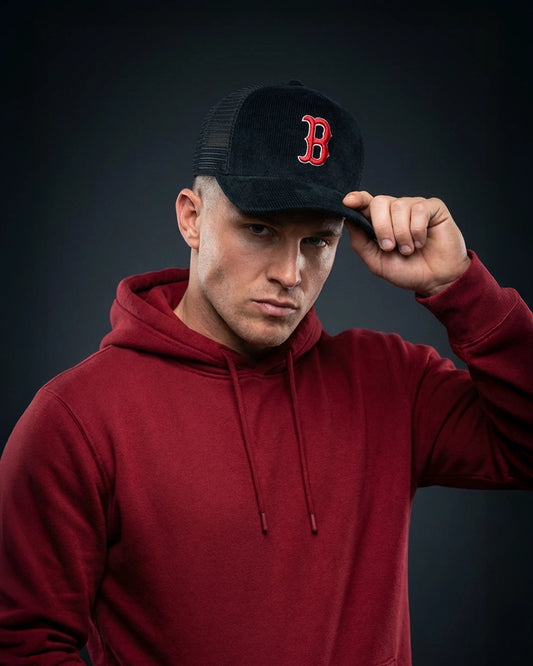 Male model wearing 999TEE Boston black trucker cap with red embroidered logo, styled with a red hoodie on a dark studio background
